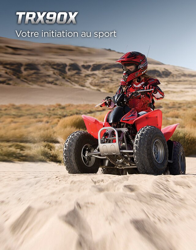 TRX90X.Initiation au sport.Image d'un jeune pilote de VTT vêtu d'une combinaison sport rouge qui manœuvre sur du terrain sablonneux inégal.