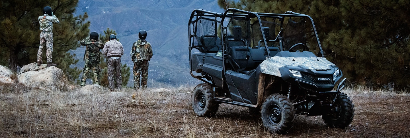 Image of four riders dressed in camoflauge looking out over a vista of rolling green hills with side-by-side parked in foreground